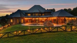 Large brick house with a dark tiled roof, warm string lights along the patio, and a wooden fence at dusk.