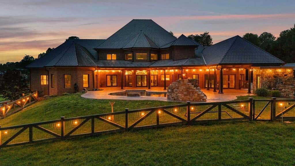 Large brick house with a dark tiled roof, warm string lights along the patio, and a wooden fence at dusk.