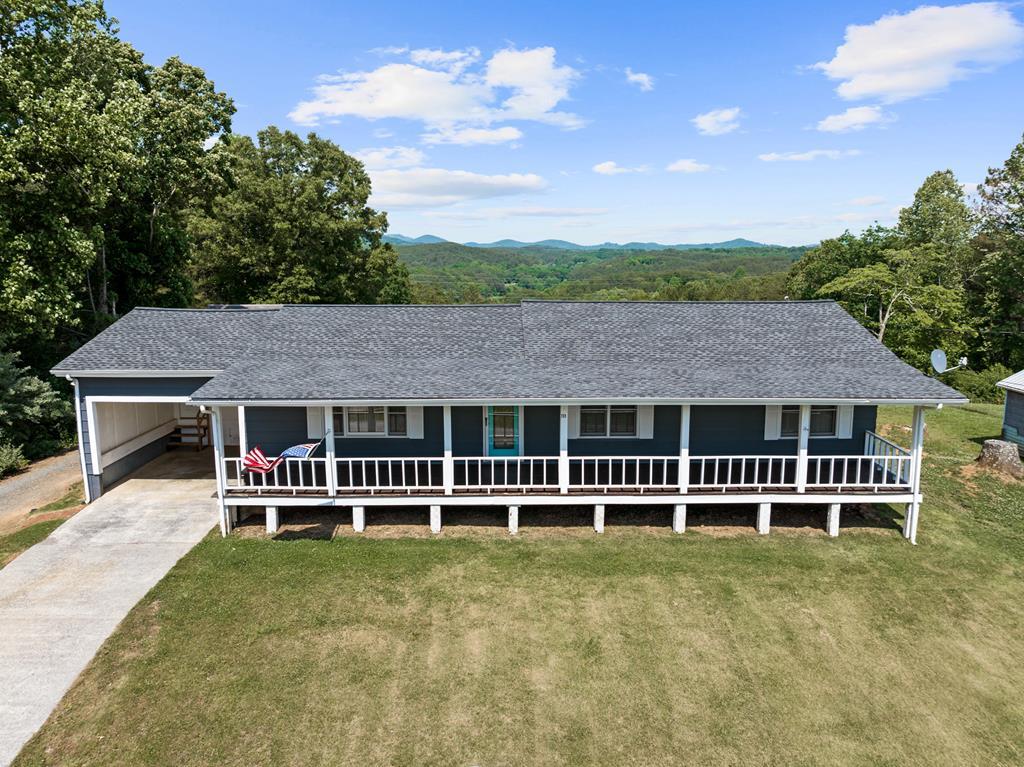 Single-story blue ranch-style house with a long white-railed front porch, set in a grassy yard with trees and distant hills on a clear day.
